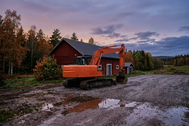 En orange grävmaskin står på en grusplan med vattenpölar framför en röd lada.